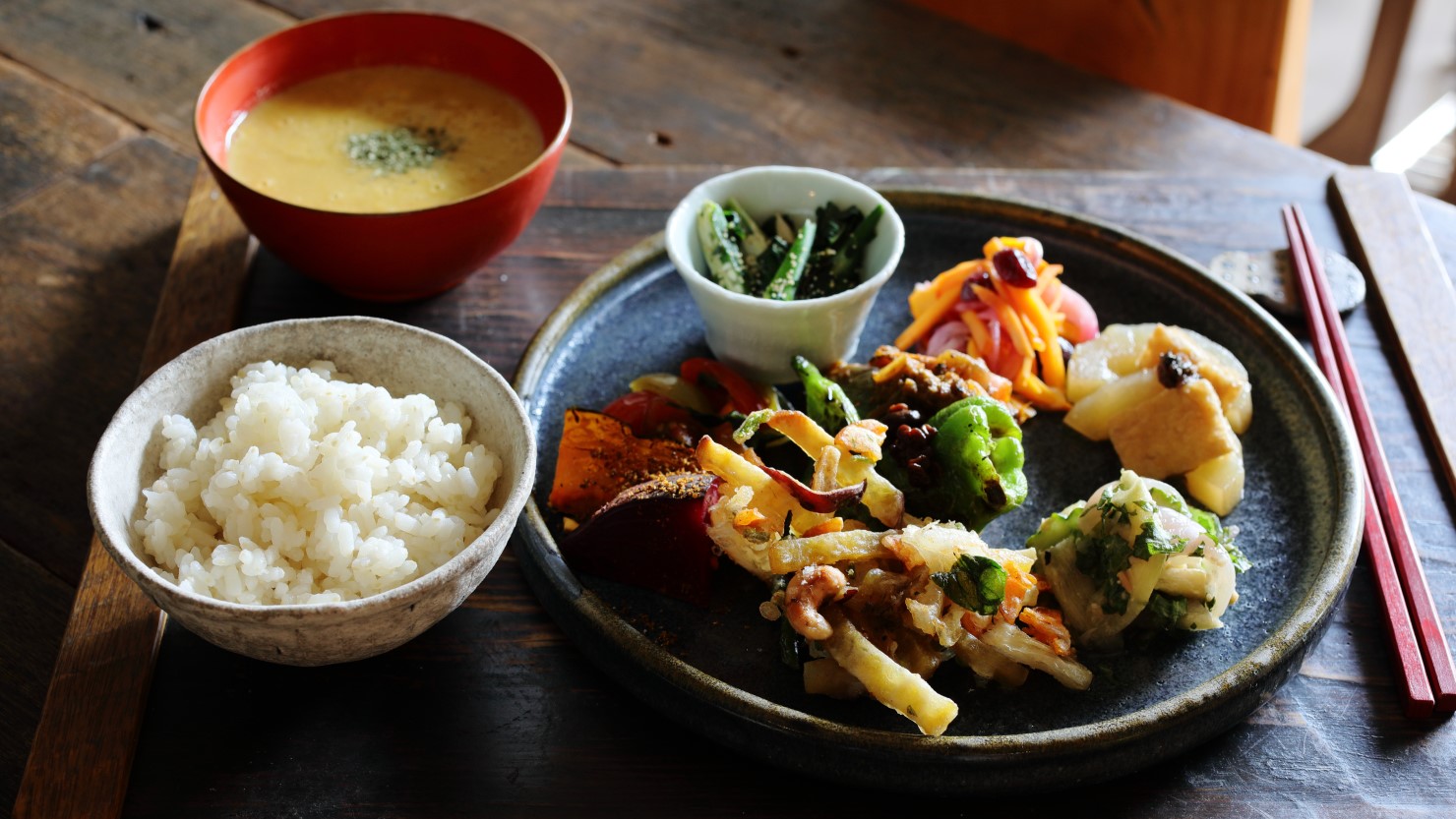 「今日の定食」(1,500円)。この日は「やさいかきあげ」「コリンキーとクランベリーのラペ」「黄瓜の煮もの」「ラタトゥイユ」「モロヘイヤとオクラのおひたし」「プチトマトのバジル炒め」「ハスイモときゅうりの白味噌あえ」「グリルやさい」「五分づきご飯」「やさいスープ」。どれも野菜の甘みなど味わいを活かしたシンプルな味付けで、変化に富んだひと皿に