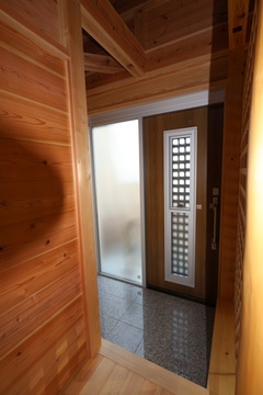 木製の壁が温もりを感じさせる玄関。シンプルなデザインで、シルバーのドアとガラスの仕切りがアクセントになっている空間。