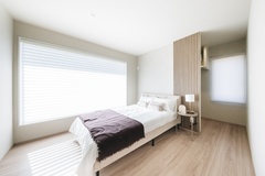 落ち着いたトーンのシンプルな寝室。大きな窓からたっぷりと自然光が差し込み、明るく開放的な雰囲気を演出している。ベッドサイドのライトが優しく空間を照らす。