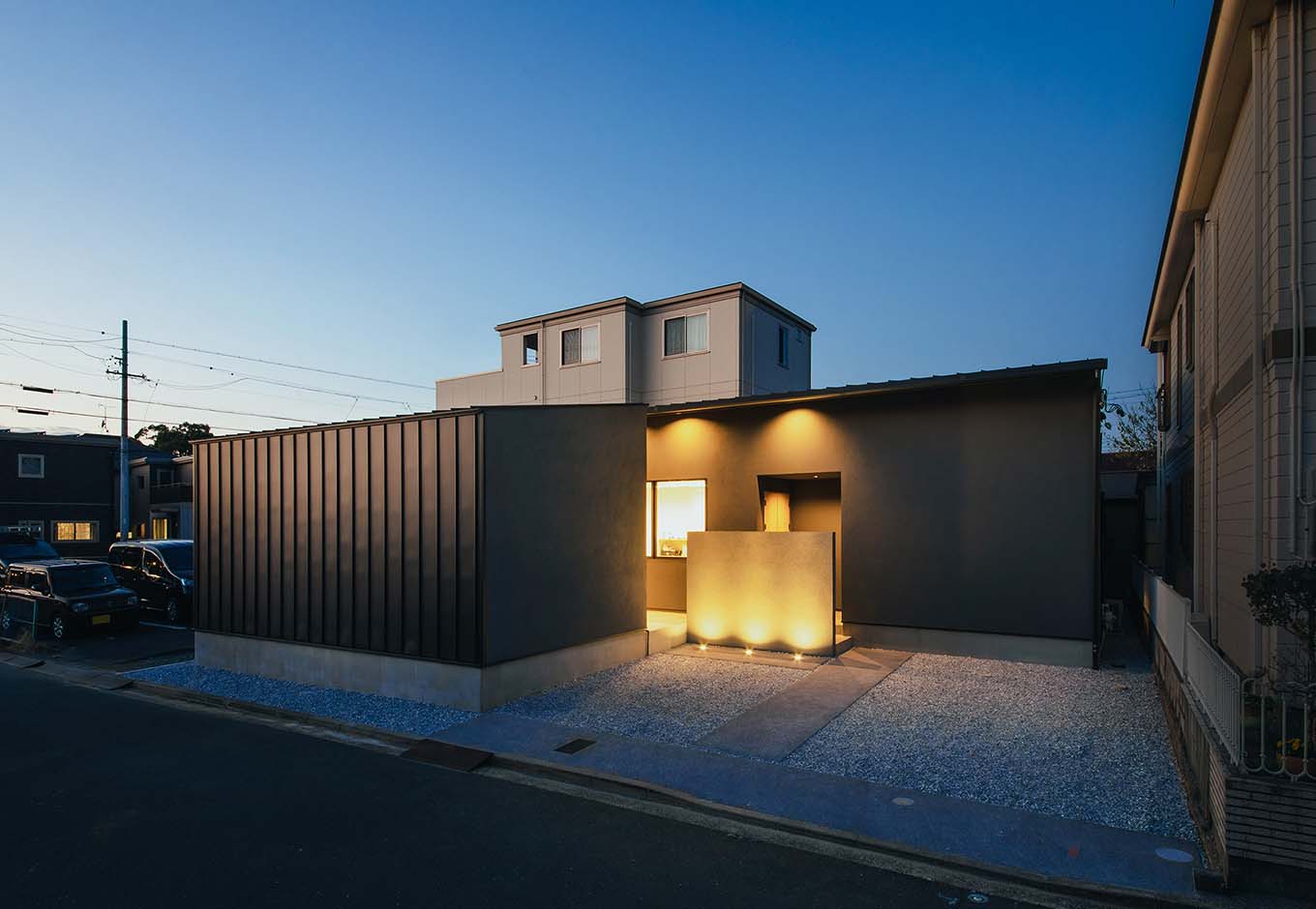 平屋建築ブルーハウス【デザイン住宅、建築家、平屋】塗り壁とガルバリウム鋼板の外壁を組み合わせ、グレーのグラデーションでまとめたシンプルモダンな外観。土地の形状に合わせてテトリス状に間取りをデザインした。夜は地面と軒天から照明を灯し、モノトーンの外観を表情ゆたかに演出