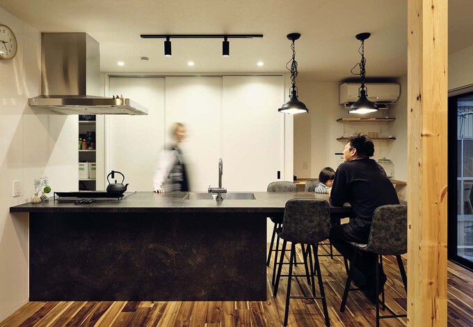 天板と扉材をブラックで統一した「GRAFTEKT（グラフテクト）」社のオープンキッチンは、ダイニングテーブルと一体型。配膳や片付けがスムーズで、家族と会話しながら作業ができる。キッチン背面には、急な来客時にも便利な大きな扉の「隠す収納」を採用。冷蔵庫もキッチン横のパントリーに隠して生活感を排除した