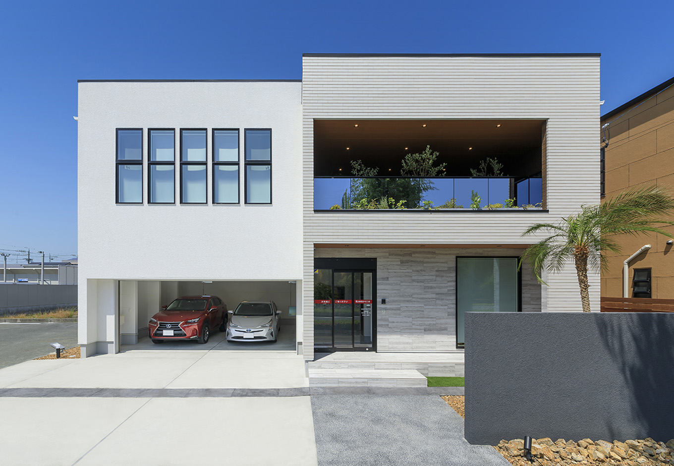 nattoku住宅【浜松市中央区新津町615-1・モデルハウス】ヤシの木、テラスバルコニーからのぞく観葉植物に、キューブ状の建物が2つ連なった外観デザインは、モナコのリゾートホテルをイメージ。白を基調としながら、のっぺりと単調に見えない理由は、随所に効かせた黒い縁取りや木目をアクセントに、装飾性のある壁やタイルでニュアンスを出しているため
