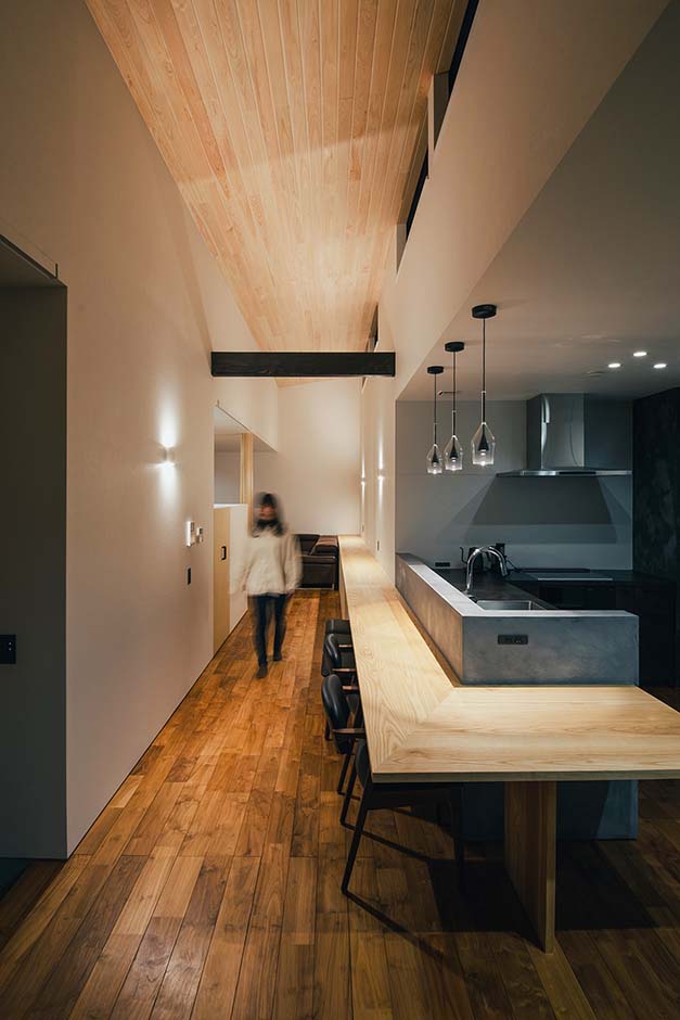 平屋建築ブルーハウス【デザイン住宅、建築家、平屋】キッチン前のカウンターはペンダント照明に照らされてバーのような雰囲気に。分けて配置したダイニングキッチンとリビングの2つの空間を、全長15mのカウンターとヒノキ板張りの高天井が穏やかにつなげている
