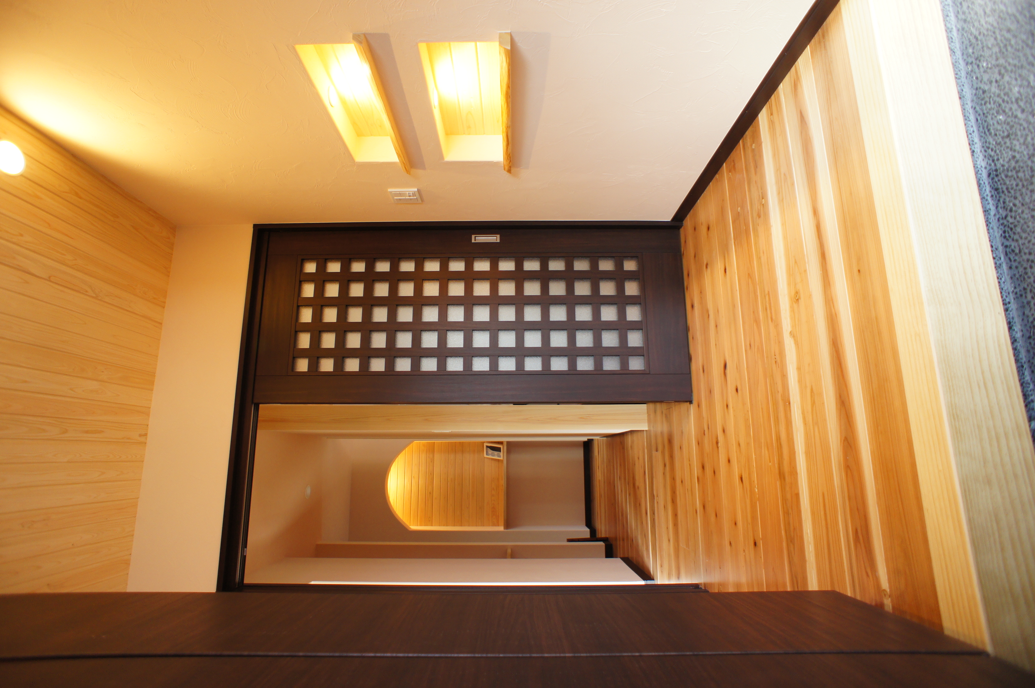 小野田産業～designers house project～｜木目調の床と暖かみのあるライティングが特徴的な玄関ホール。収納棚がシンプルかつ実用的で、洗練された印象を与えるデザインである。