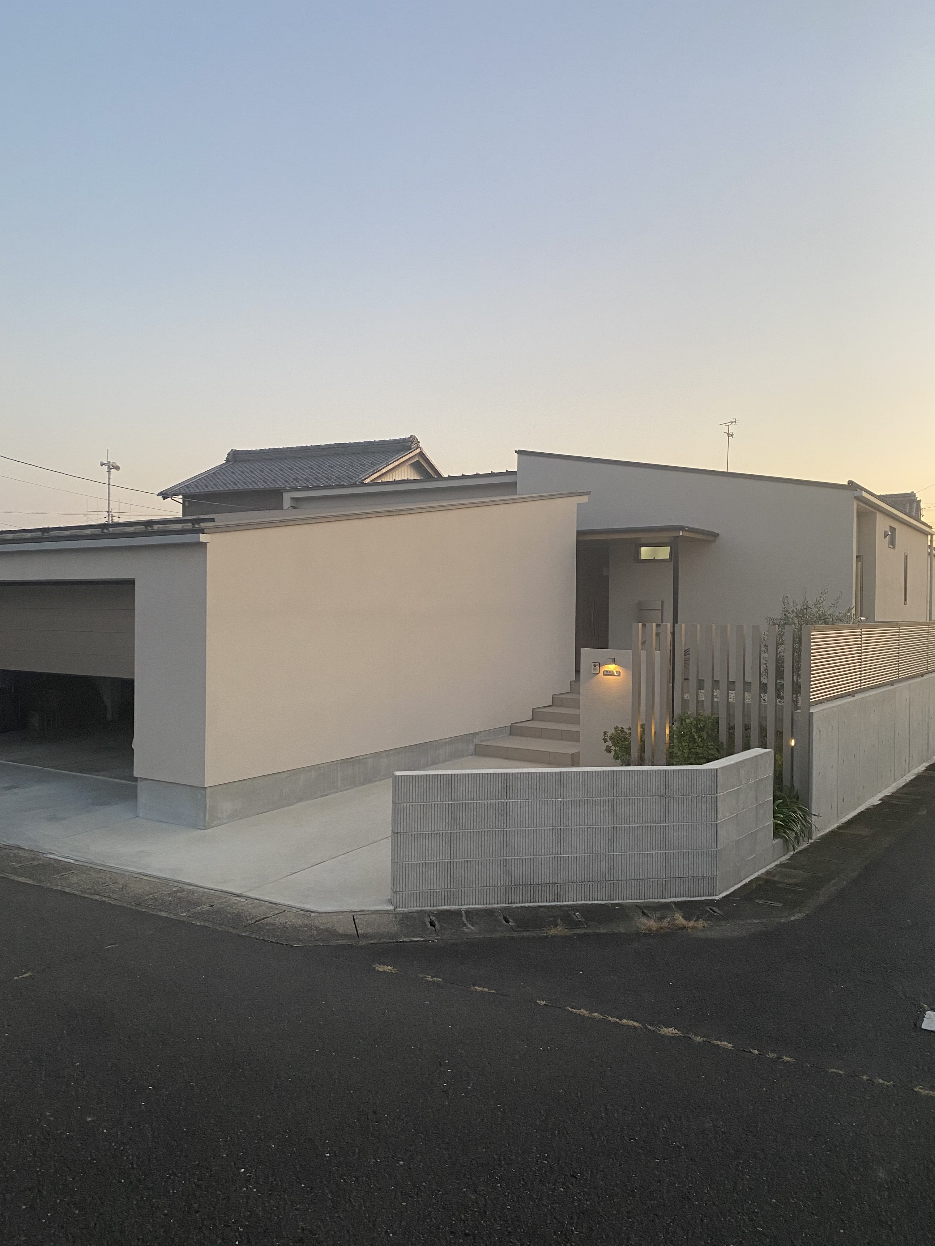 安田住宅｜安田建築設計室｜シンプルでモダンなデザインの平屋外観、ガレージシャッターを動かしている様子。柔らかな夕暮れに照らされるシックな色合いと、プライバシーを重視した構造が特徴である。