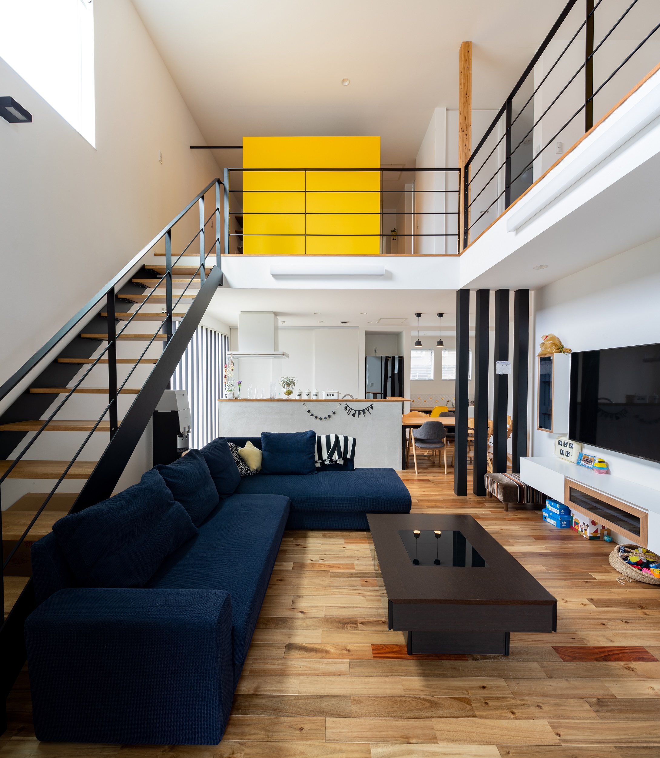 S.CONNECT（エスコネクト）【デザイン住宅、子育て、建築家】LDKは、大きな吹き抜けとアイアンのストリップ階段を採用し、開放的な空間に。ナチュラルな無垢の床にアイアンの階段や柱の黒でメリハリをつけた。2階にあるイエローのアクセントウォールもインテリアの一部に