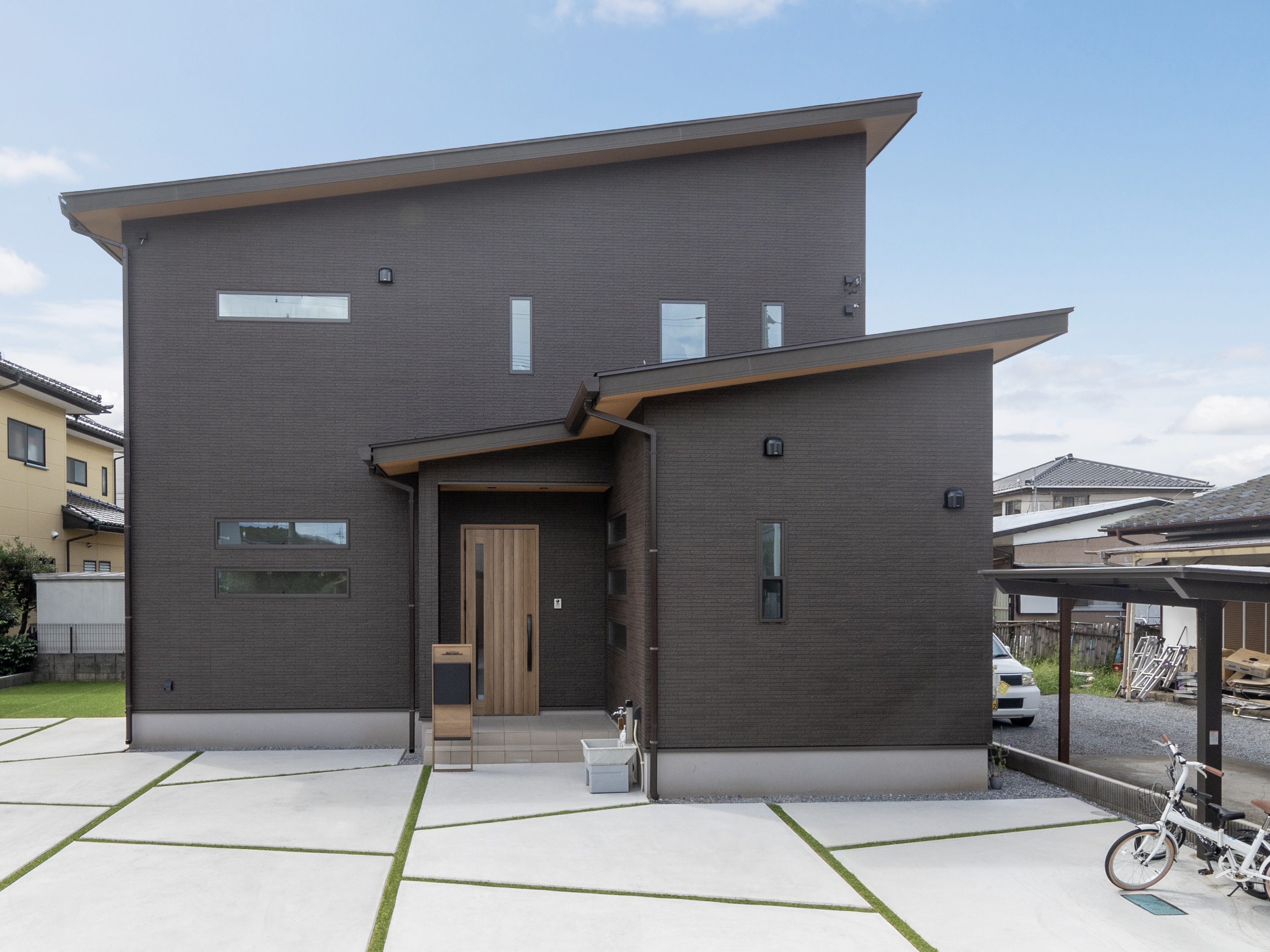 エスホーム｜下屋(げや)と呼ばれる１階部分も合わせて片流れにした屋根。外壁の色はスペルトブラウン。エアコンの室外機と配管が前面にこないように配慮。「あみだくじ模様」のカーポートは遊び心がある。