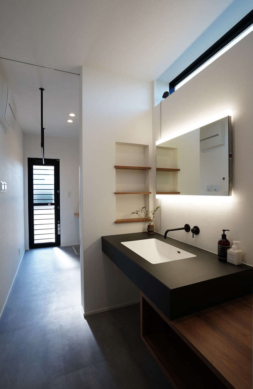 アールスタジオ【デザイン住宅、平屋、インテリア】造作の洗面スペースも、カウンターに黒を採用。鏡の背面に仕込んだ間接照明が、ホテルライクな雰囲気を醸し出す。洗面スペースの奥にはランドリーと勝手口が設けてあるため、室内干しも外干しもできて便利
