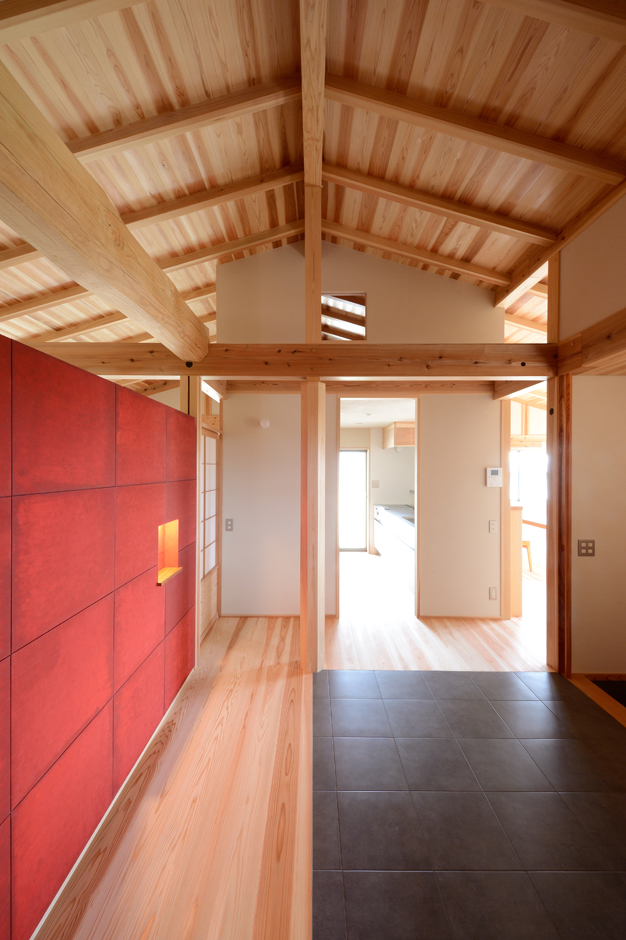 石牧建築【デザイン住宅、二世帯住宅、建築家】玄関から室内に入った途端、太いマツの太鼓梁と赤い漆塗りのアクセントウォールが目に飛び込んでくる。玄関ホールは南面で日当たりが良いため、フロアタイル貼りの「土間リビング」として利用。書棚も設けてあり、お父さまが読書をゆったり楽しめる