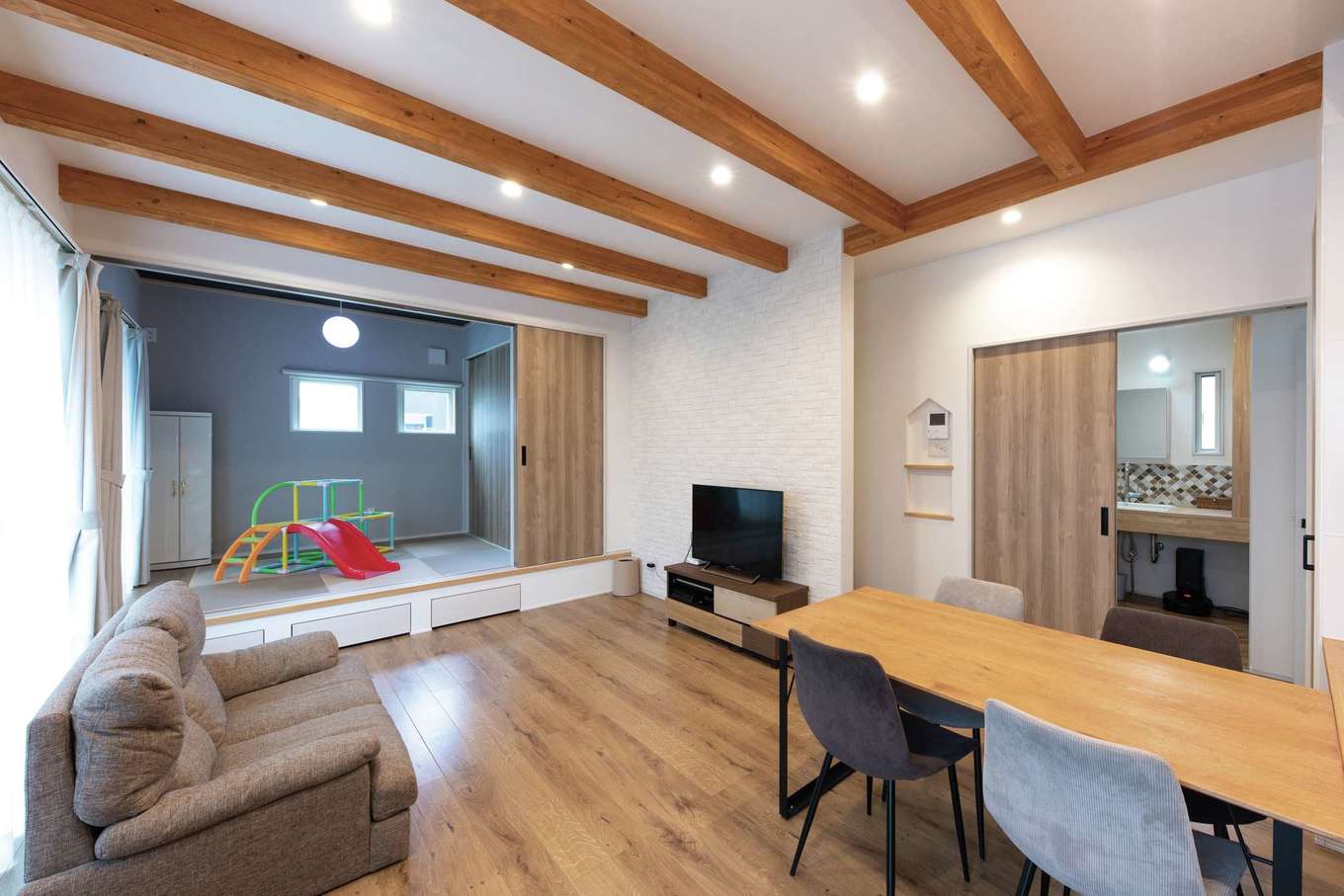 小玉建設【デザイン住宅、子育て、間取り】19.5畳のLDKに3.8畳の和室が連続。普段は開いて広さを演出しつつ、遊び場やお昼寝スペースとして活躍。来客時は閉じて客間となる。洗面室の左手に階段。音やにおいが2階にいかないように空間を区切りながら、2階に上がるときにはキッチンの横を通るようにした。モニターニッチも山型に