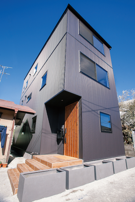 建築システム（狭小住宅専門店）【1000万円台、デザイン住宅、インテリア】ブラックメタリック色のガルバリウム鋼板。この無機質な冷ややかさとウッド調のポーチ・玄関ドアの組み合わせが絶妙なバランスの外観を醸 し出す。天候や時間帯で表情が変わる不思議な建物だ。キュービックスタイルの中はどうなっているのか、興味をそそられる