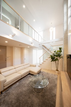 吹抜けのある広々としたリビング。白いソファとガラスのテーブルが洗練された雰囲気を醸し出している。階段は透明なガラス手すりでスタイリッシュ。