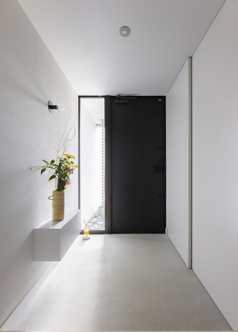 白とグレーで統一され、静けさと上品さが漂う玄関。大きな白い扉の奥はシューズクローク。壁の反対側はリビングの壁掛けテレビのスペースで、機材の配線を隠してしまうボックス・スペースを玄関に突出させ、飾り棚として利用している