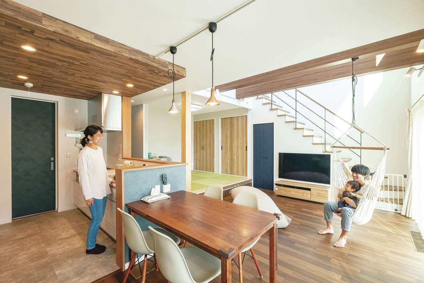 イデキョウホーム【省エネ、間取り、屋上バルコニー】吹き抜けからの光に包まれるLDK。細かな間取りを伝えたわけではなく、「キッチンから家族が見えるようにしたい」「子どもが大きくなっても顔をあわせるように」「ハンモックでゆらゆらできたら」といった要望を設計士が形にしてくれた。素材や建具の色で迷ったときは、インテリアコーディネーターに相談。開放感に落ち着きと楽しさが溶けあう空間に仕上がった。右手には広々としたウッドデッキも用意されている
