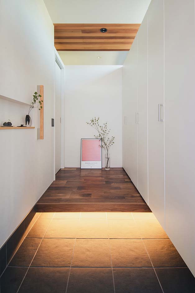 平屋建築ブルーハウス【デザイン住宅、建築家、平屋】間接照明がタイルの土間をやさしく照らす玄関。土間からホールまでの壁は一面シューズボックスに。内装は白で統一し、壁面に絵画を飾るなど、インテリア小物で空間を彩っている。周囲の壁の上部をオープンにすることで、明るさと開放感をもたらした