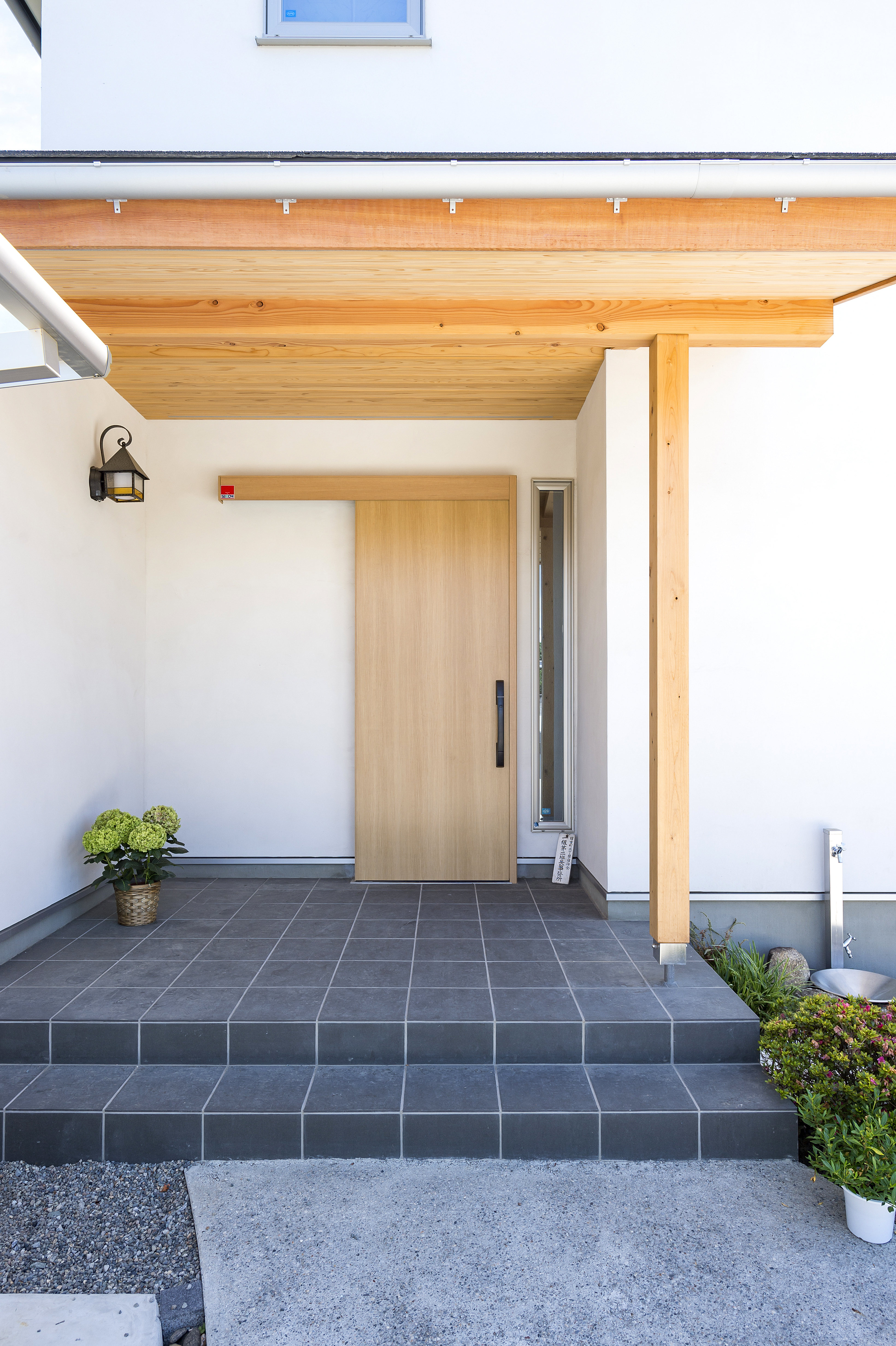 しあわせの住まい一級建築士事務所｜ナチュラルな木材の軒とガラス入りの木製ドアが印象的な玄関。訪れる人を優しく迎え入れるデザインで、玄関前には鉢植えやグリーンが配置されている。
