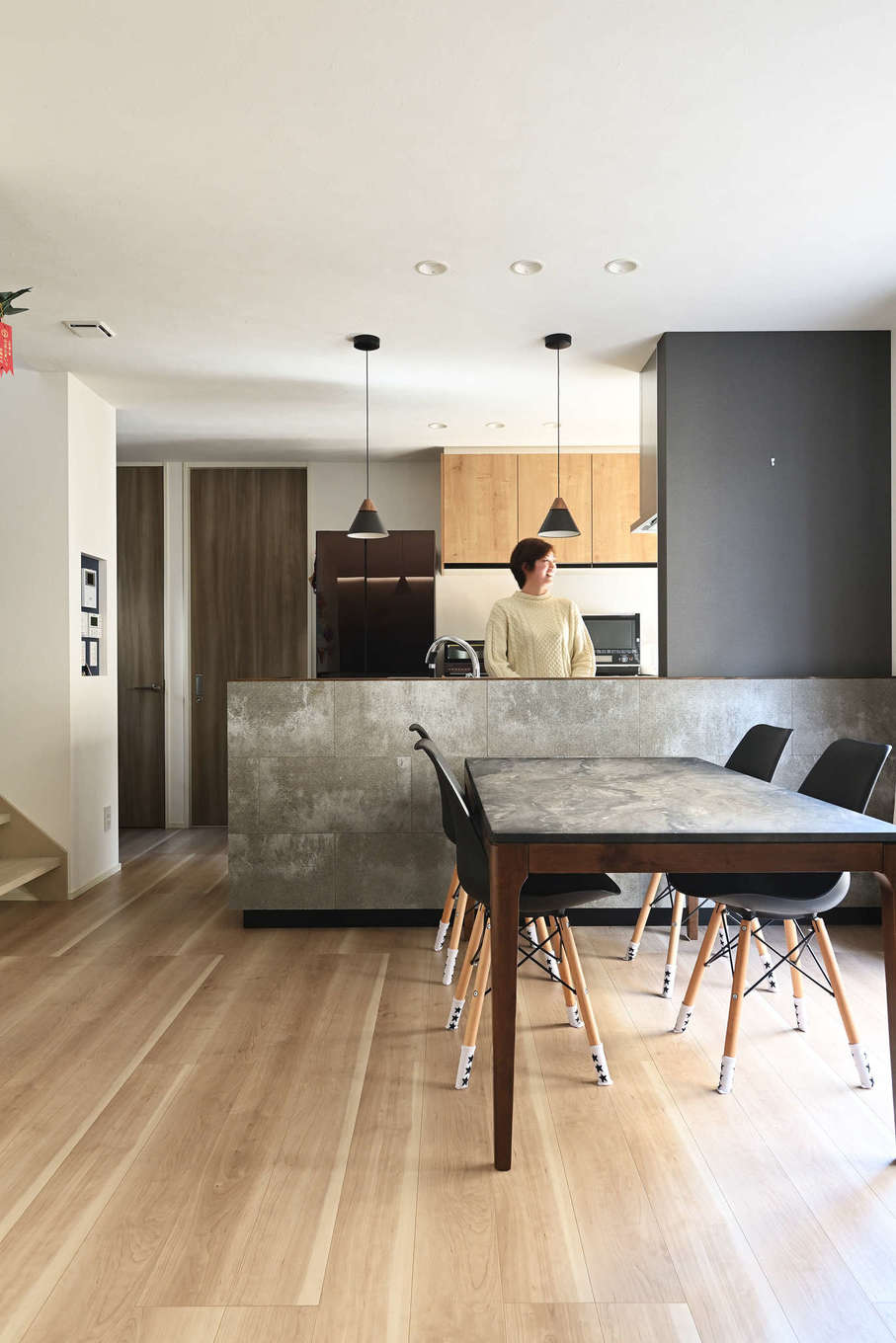 トーリンホーム【デザイン住宅、子育て、間取り】リビングのアクセントクロスと同様に、対面キッチンの腰壁にも家具調に「ソリド」を張って統一感を演出。ダークグレーのクロスやペンダント照明とも絶妙にマッチして、スタイリッシュな空間に仕上がった。ダイニングテーブルもデザインテイストを揃えた