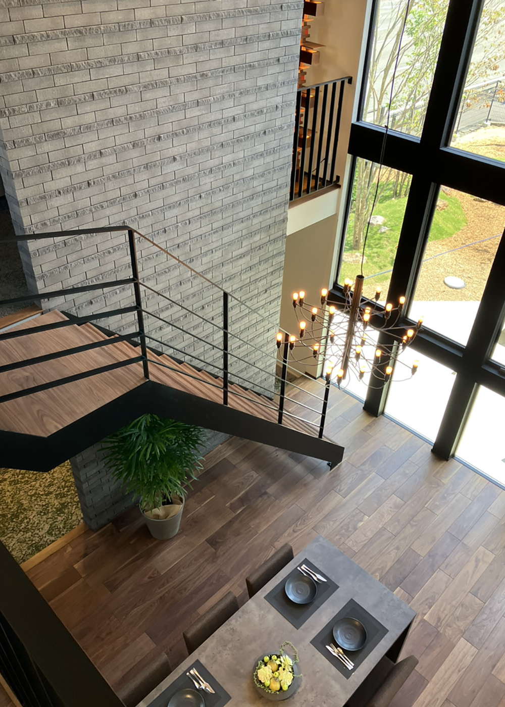 マルモホーム 建設のマルモ【静岡市駿河区桃園町1-1・モデルハウス】ダイナミックな吹抜けに天井まで届く連窓、リビングから2階へ続く鉄骨階段が、立体的な視覚効果を創出。抜け感と格調高い雰囲気を添えている。床はモノトーンの内装に合うダーク寄りのウォールナットを採用