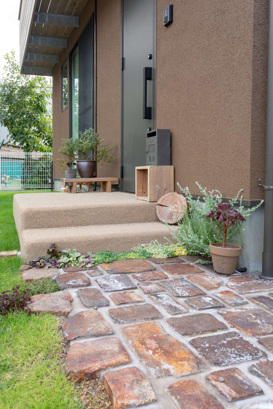 フジモクの家（富士木材）【子育て、自然素材、間取り】絵本の世界から抜け出してきたような、大人かわいい雰囲気のアプローチ。グリーンが映えるレンガ敷きの小径、角を丸くした洗い出しのポーチなど、モカブラウンの塗り壁に合わせてコーディネート