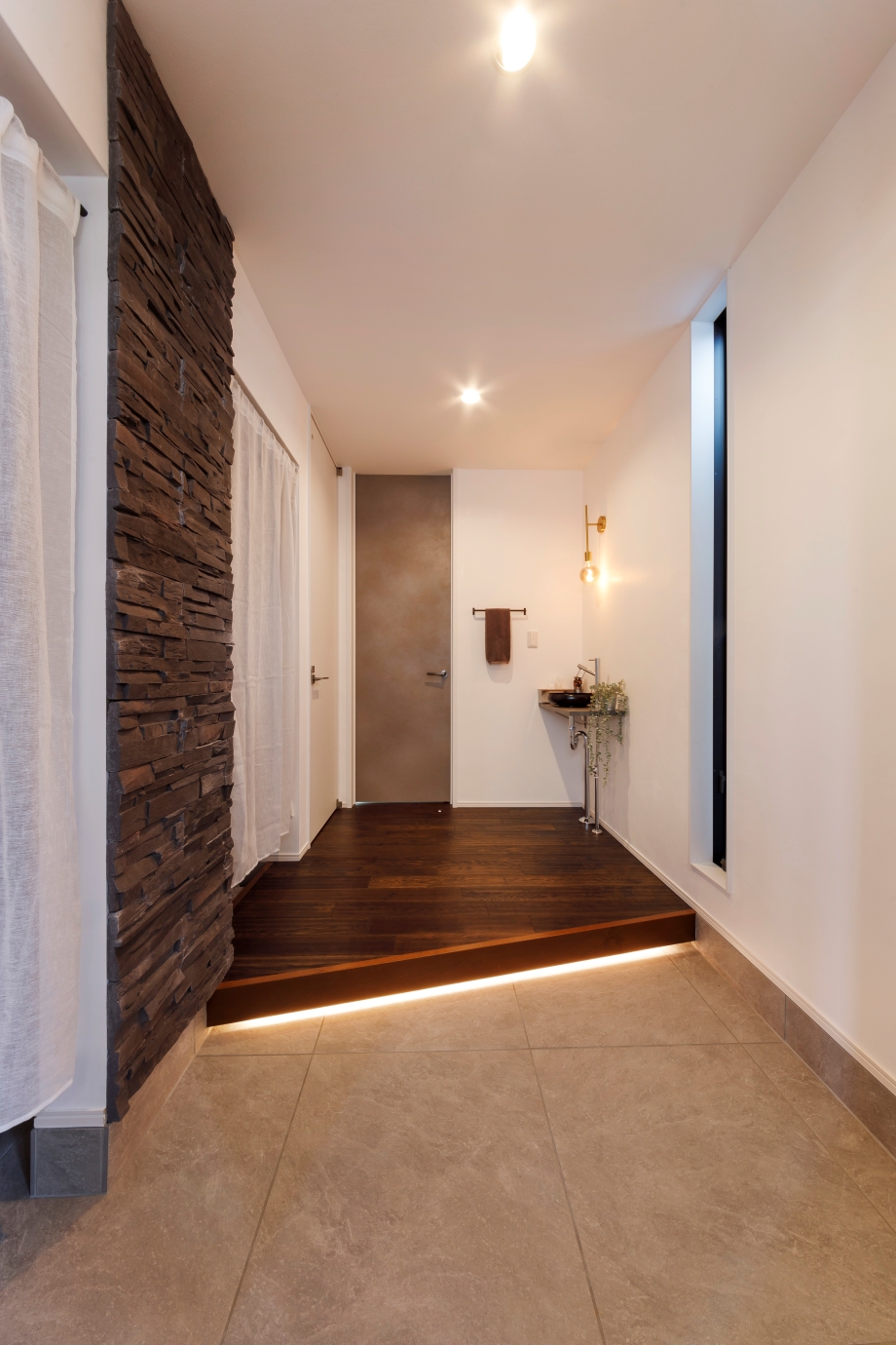 nattoku住宅【デザイン住宅、子育て、間取り】玄関はストーンタイル、間接照明、マットな質感の建具などにより、エレガントな仕上がりに。通り抜け可能なシューズクロークには可動棚やコート掛けのほか、サーフボードを収納するスペースも完備。「幼少期から自分の命を守る意識付けになれば」というご主人の要望で、玄関ドアの上には、非常口であることを示すグリーンの誘導灯を設置している