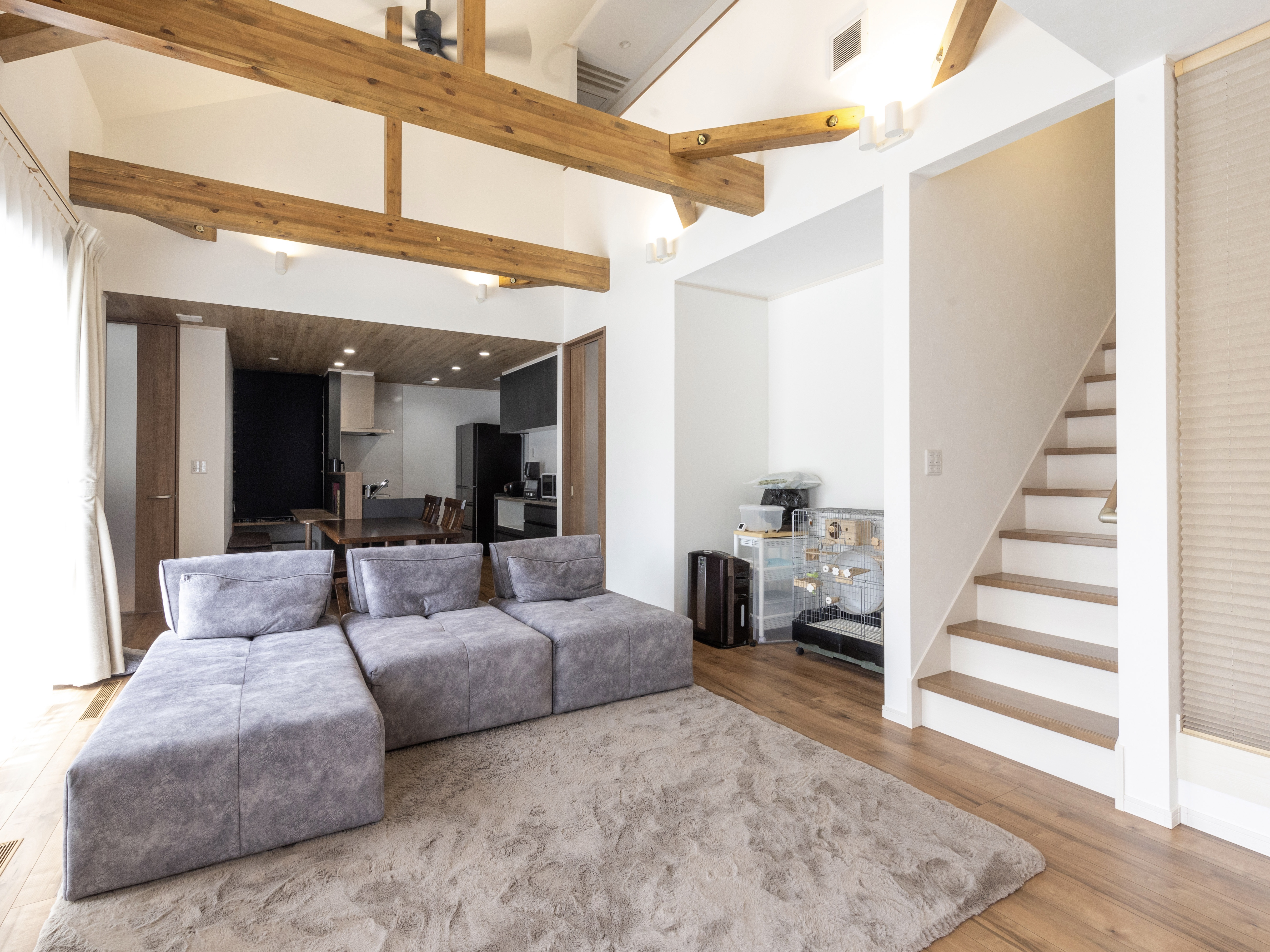 エスホーム｜落ち着いた色合いのソファが配置されたリビング空間。高い天井の梁がアクセントとなり、開放感を演出している。右側には階段があり、多目的な使い方が可能である。