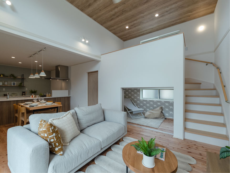 ヘスタホーム｜木目調の勾配天井が特徴的な、明るく開放的なリビング。大きな窓と白壁が空間を広々と見せ、ゆったりとしたソファがくつろぎを提供。ダイニングキッチンとの一体設計で、家族の団らんを促す温かな空間です。