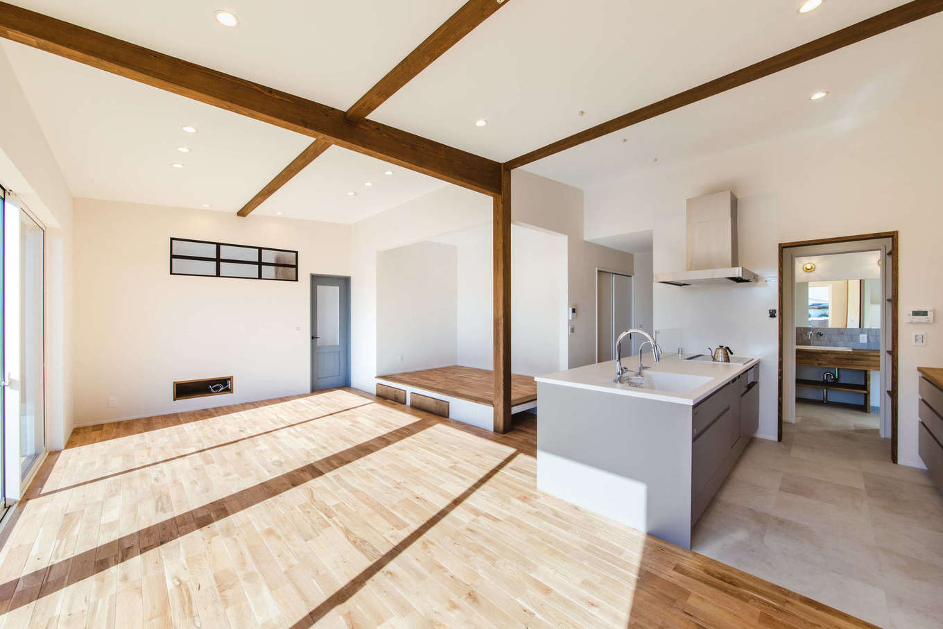 Select工房【デザイン住宅、間取り、平屋】風が抜けるLDKは、小上がりのヌックスペースを加えれば21畳と広々。壁は塗り壁仕上げ。壁掛けテレビの配線を隠すために、リビングの壁の一部に空配管を通している。オークの床や勾配天井に配した化粧梁など木の素材感を楽しめる仕上げに