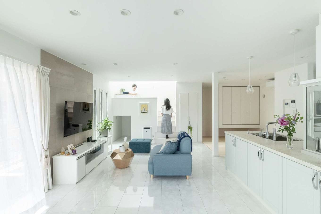 HAPPY HOUSE（山田工務店）【デザイン住宅、間取り、インテリア】ハワイやヨーロッパが好きな奥さまの希望で、LDKは洗練とリゾート感が溶け合う空間に。大理石調のフローリングにあわせ、全体が品のあるホワイトでまとめられた。右奥の畳コーナーも、あえて段差をつけず、LDKとの一体感を優先。テレビ背後にはアクセントとしてエコカラットが採用され、ほどよい落ち着きが添えられている。掃除のしやすさも大切なポイント。適所に十分な量の収納が用意されていて、常にキレイな状態が保たれる