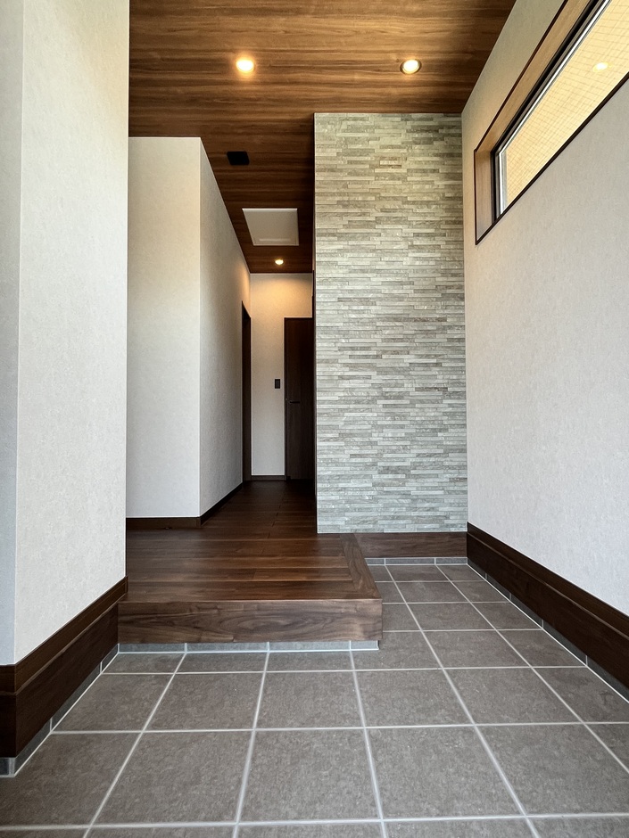 シックなタイルフロアが印象的な玄関。壁のアクセントに用いた石材調のデザインが空間を引き締め、来訪者を迎え入れる。