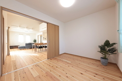 木製フローリングが広がる明るい居室。大きな窓から自然光が差し込み、開放感を演出している。隣接するリビングが見渡せるため、家族の気配を感じながら過ごせる空間。