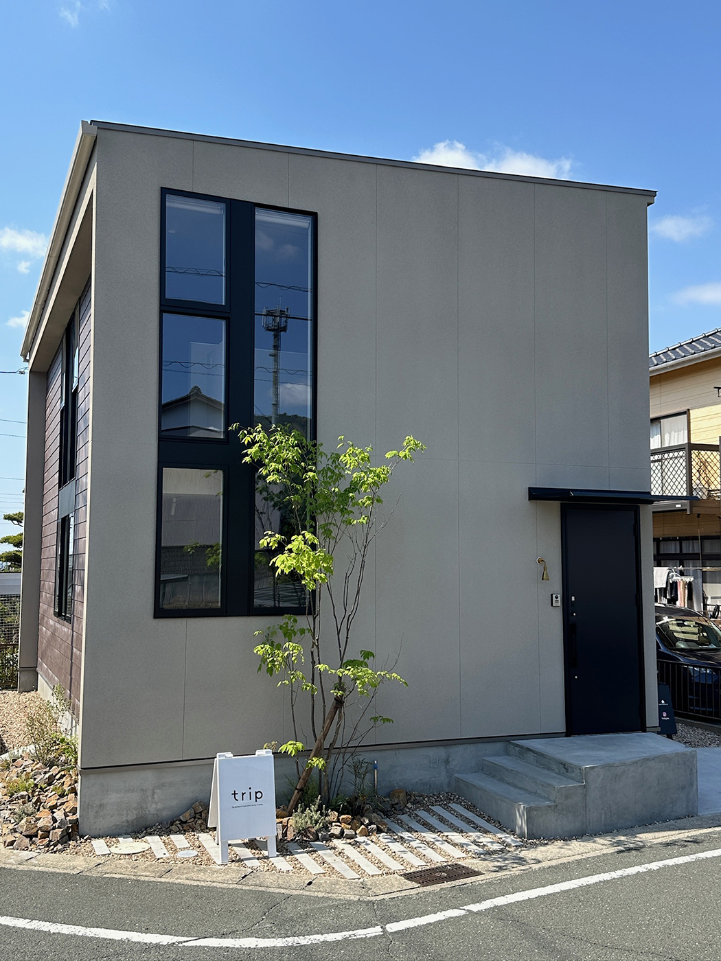 BASE.trip【デザイン住宅、間取り、インテリア】総二階建てのスタイリッシュなグレーの外観デザイン。アイボリーの塗り壁に黒い窓枠のサッシが美しく調和している。シンボルツリーはアオダモで、建物の雰囲気に合わせて外構もトータルにデザインする