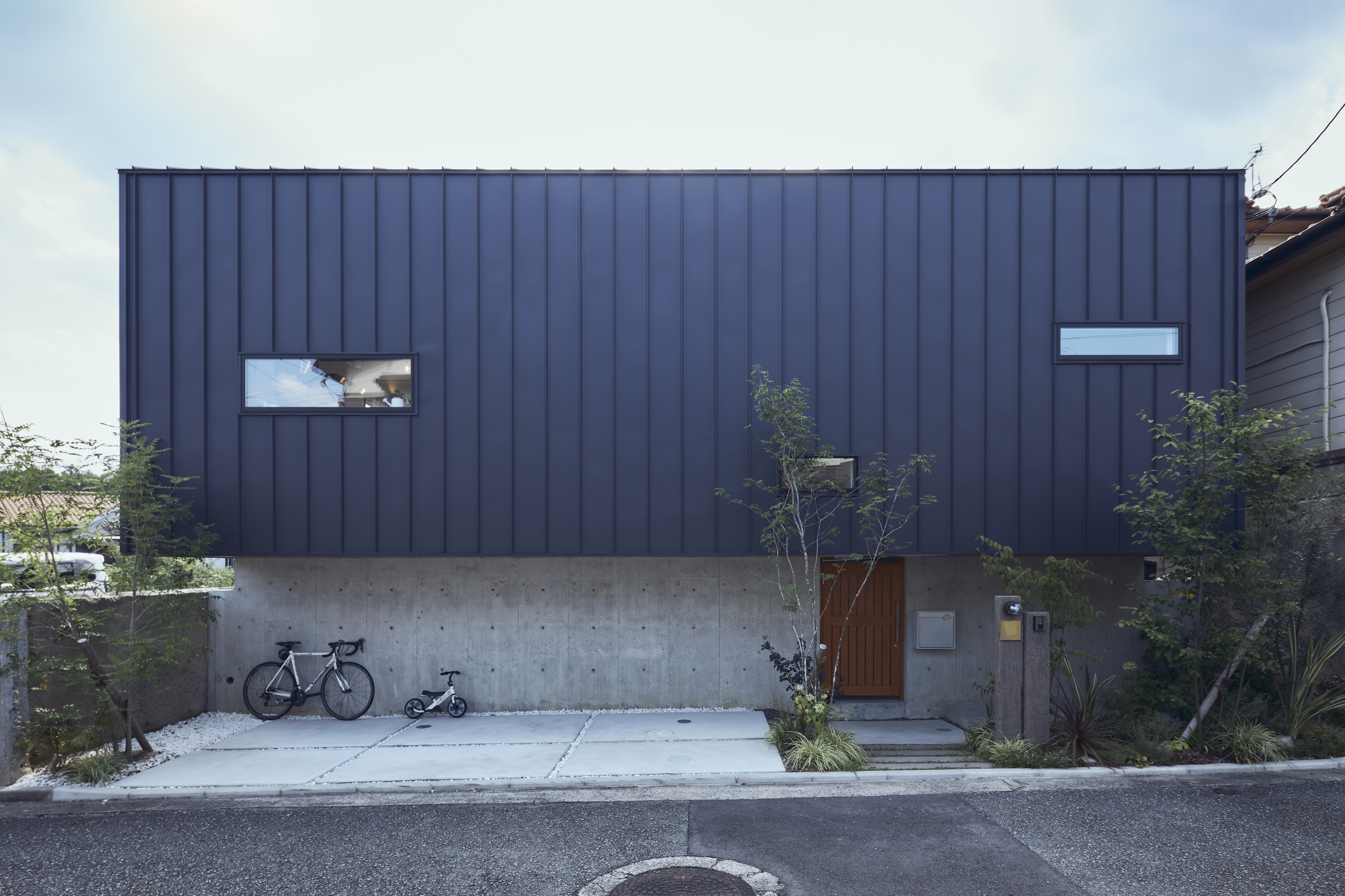 建築設計事務所 KADeL｜キャンチレバーを取り入れ、建物の上部を前面に。コンクリートの壁や屋根から続くガルバリウム鋼板、木製の引き戸など、素材をバランスよく組み合わせたデザインに青々と茂る植栽がしっくり。建築的な美しさにこだわったファサードになっている
