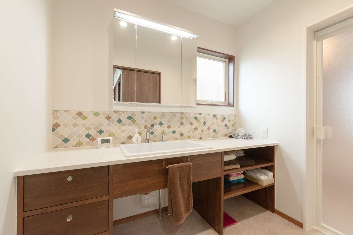 ホームプラザ大東【デザイン住宅、子育て、収納力】タイルでかわいさを添えた造作洗面台は、朝の準備で渋滞しないよう幅が広く取られた。流しも医療用の大きなサイズのものが選ばれている。左手には室内物干しとアイロン掛け用のカウンターが備えられた家事室。浴室から外の物干し場までが一直線で結ばれ、洗濯の効率を高めている