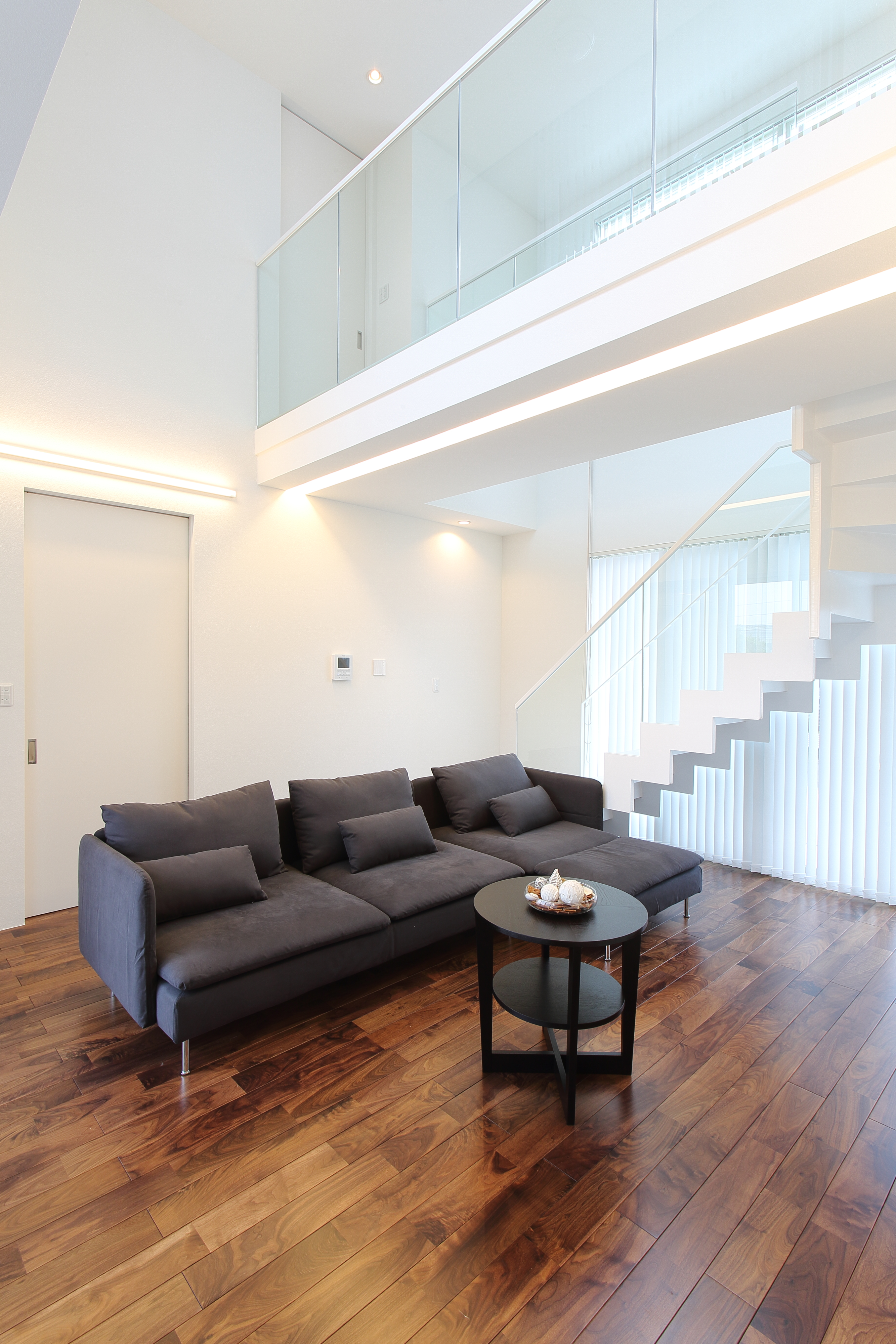 丸昇彦坂建設【デザイン住宅、間取り、ガレージ】大きな窓から見える外の景色と吹抜けの天井で、開放感を強調したリビング。白い空間にウォールナット材の床を組み合わせて、落ち着きのある雰囲気を演出