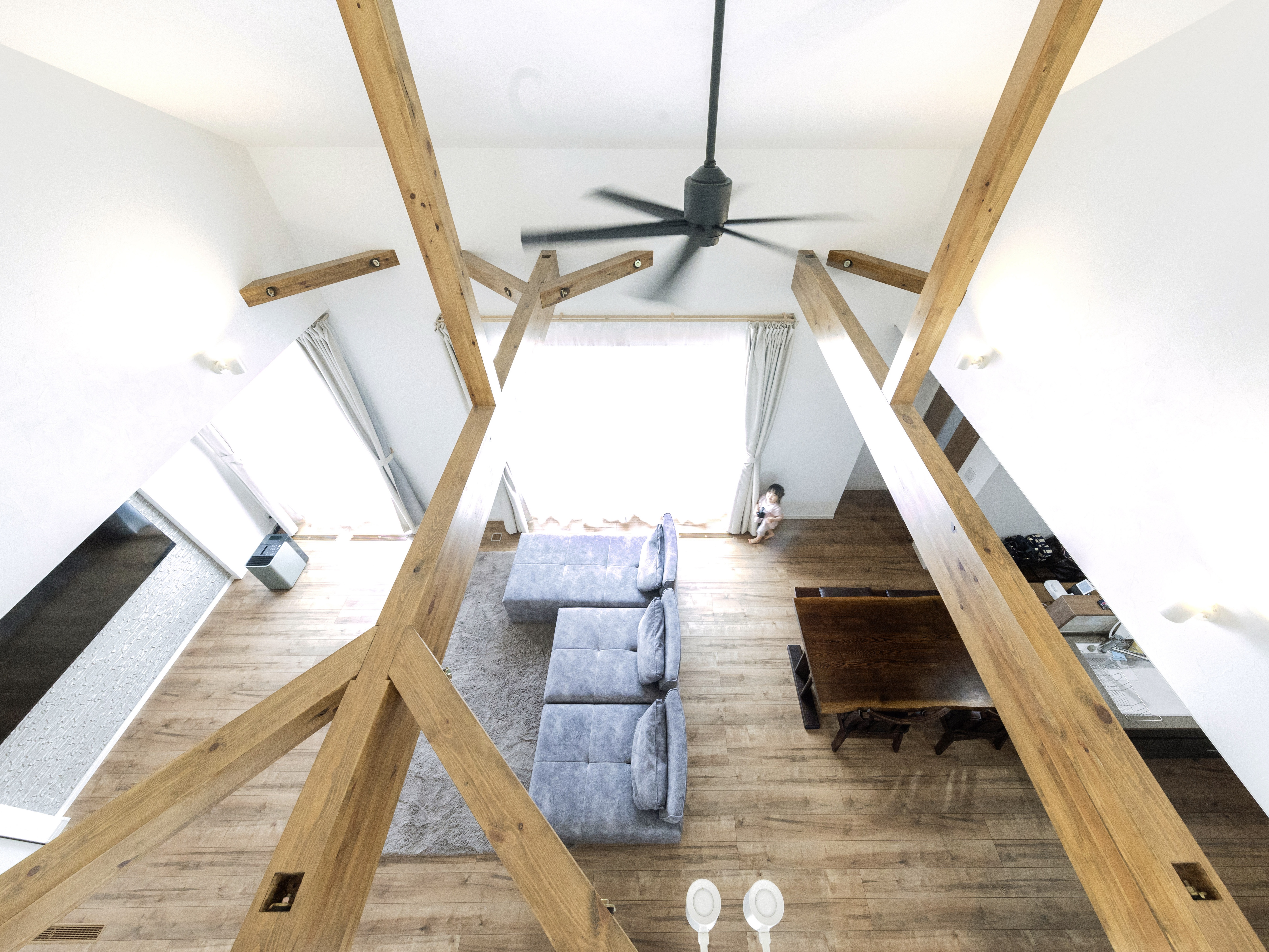 エスホーム｜開放感のある吹き抜けのリビングダイニング。木の梁と大きな窓が、明るく広々とした空間を演出している。天井のファンが空間のアクセントとなっている。