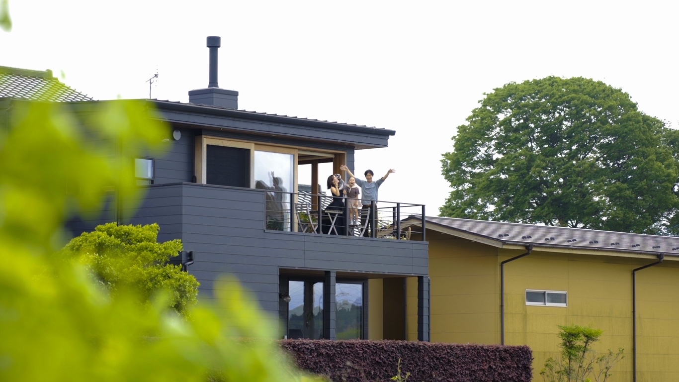 伸興建設｜モダンな外観の二階建て住宅。バルコニーから景色を楽しむ家族の姿が見受けられる。シックな色合いが際立つおしゃれなデザインである。