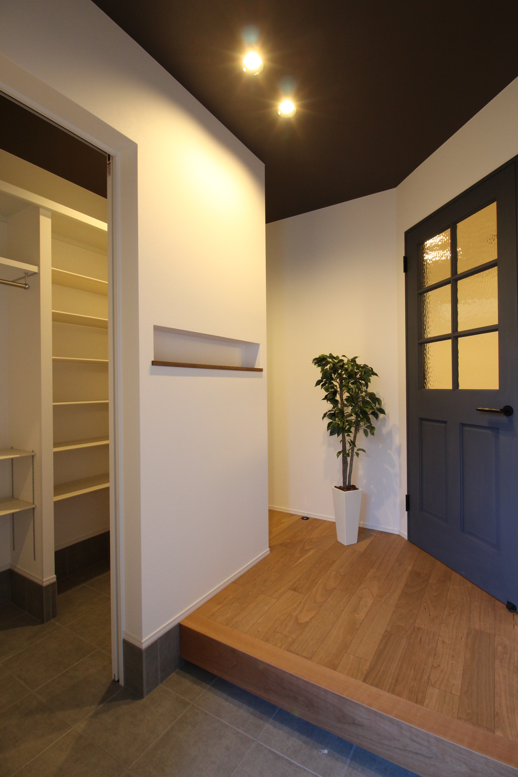 小野田産業～designers house project～｜シンプルで直線的なデザインの玄関。照明と観葉植物が柔らかい雰囲気を演出し、扉の向こうの空間への期待感を高めている。
