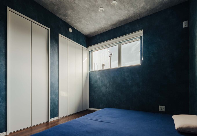 深いブルーの壁とグレーの天井が目を引く、約6畳の主寝室。ローベッドでも使いやすいようコンセントを低い位置に設置した。シーツは壁紙に合わせてコーディネート