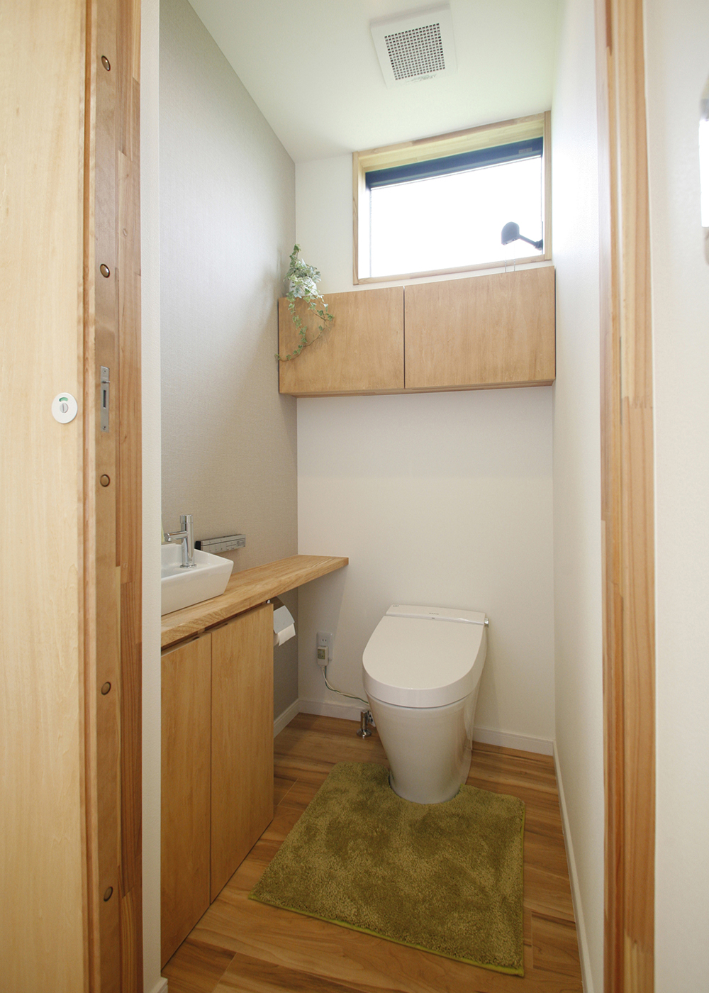 タツホーム・竜建｜コンパクトながら機能的なトイレスペース。木目調の収納とグリーンのアクセントが、清潔感と落ち着きを与える。