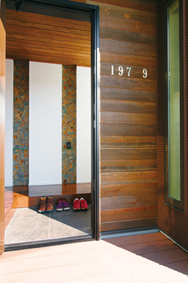 フロリダの邸宅を思わせる、アプローチから玄関へ続く長いウッドデッキ。アメリカに2年間滞在していたご主人。奥さまの要望で、当時の家のように玄関ドアの脇に番地を示す大きなプレートを貼った。完成見学会でも「真似したい！」と大反響を呼んだ