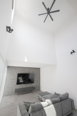 シンプルでモダンなリビング。高い天井と大きな壁掛けテレビが印象的で、快適なリラックス空間を提供する。灰色のソファが落ち着いた雰囲気を演出している。