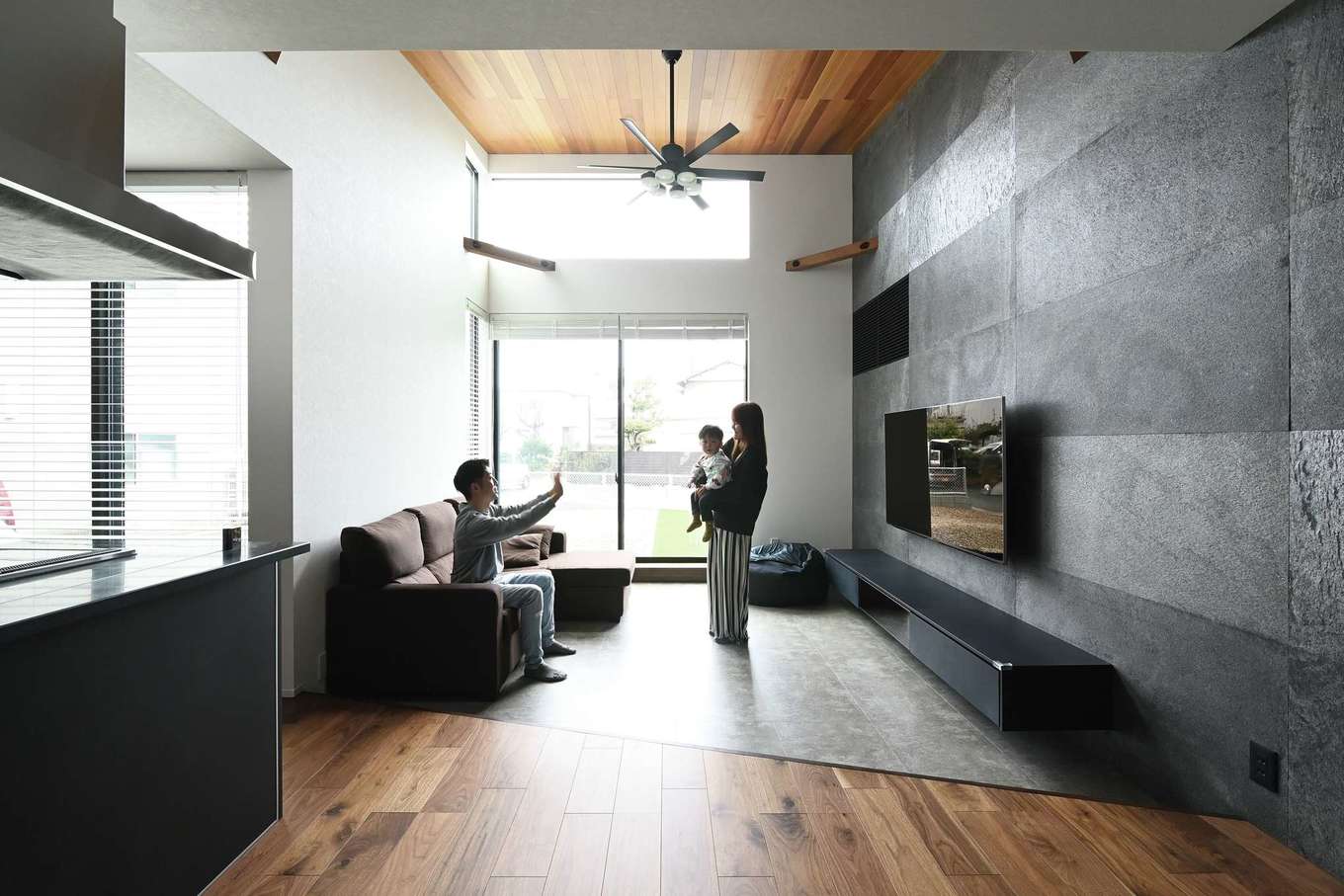 REIWA HOUSE（令和ハウス）【デザイン住宅、間取り、平屋】23畳のLDK。テレビステーションの壁全面に天然石を張り、高級感を演出。リビングをフロアダウンさせ、さらにレッドシダーの勾配天井も採用したことで、いっそうの開放感が生まれた。大きな窓越しに緑の庭が見える