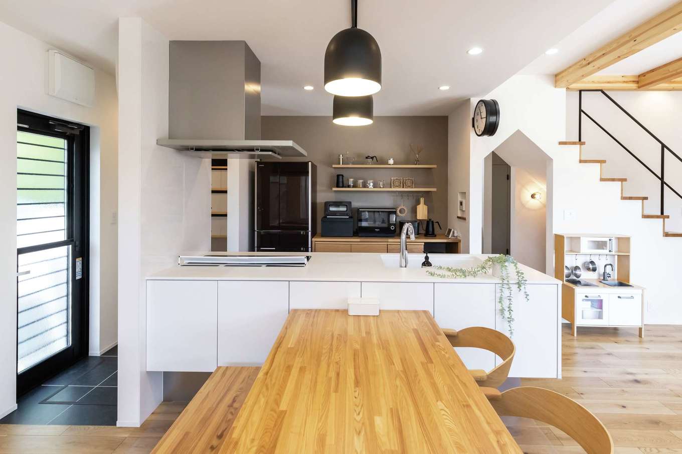 SEVEN HOUSE/セブンハウス【デザイン住宅、間取り、平屋】将来的にインテリアの好みが変わっても、空間の色に合わせやすいよう、キッチンは白を選択。ご主人も料理をするので、ワークスペースを広めに取った。ぐるぐると回遊できるアイランドキッチンは、家事効率をグーンと高めてくれる。カップボードは飾り棚を造ってもらい、見せる収納に。大きなダイニングテーブルは同社の西岡さんが組み立ててくれた