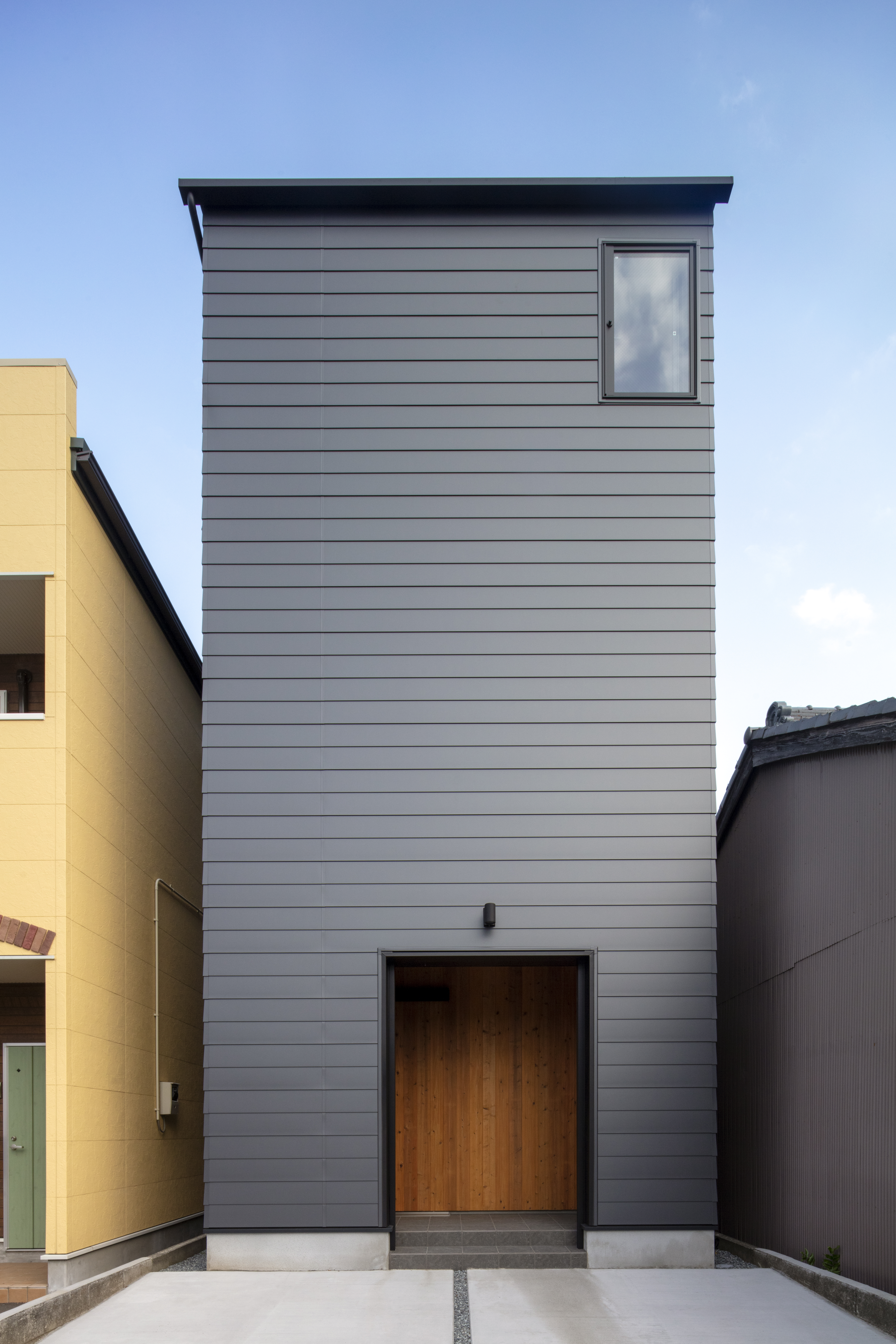 一級建築士事務所 GO_AN (ゴアン)【デザイン住宅、二世帯住宅、狭小住宅】大通りに面した北側は窓を少なめにして、外からの視線をカット。ガルバリウム鋼板の外壁が醸すインダストリアルな雰囲気に、玄関ドアの木目がやわらかなアクセントを添える。玄関前にはご主人の趣味の車を2台停められるスペースも確保
