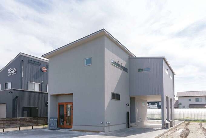淡いグレーを基調にした外観。正面は塗り壁にして木扉を付け、店舗の入り口に。縦長の敷地を有効活用し、西面にピロティ付きの自宅用玄関を設置。奥にも駐車場があり通り抜けできる