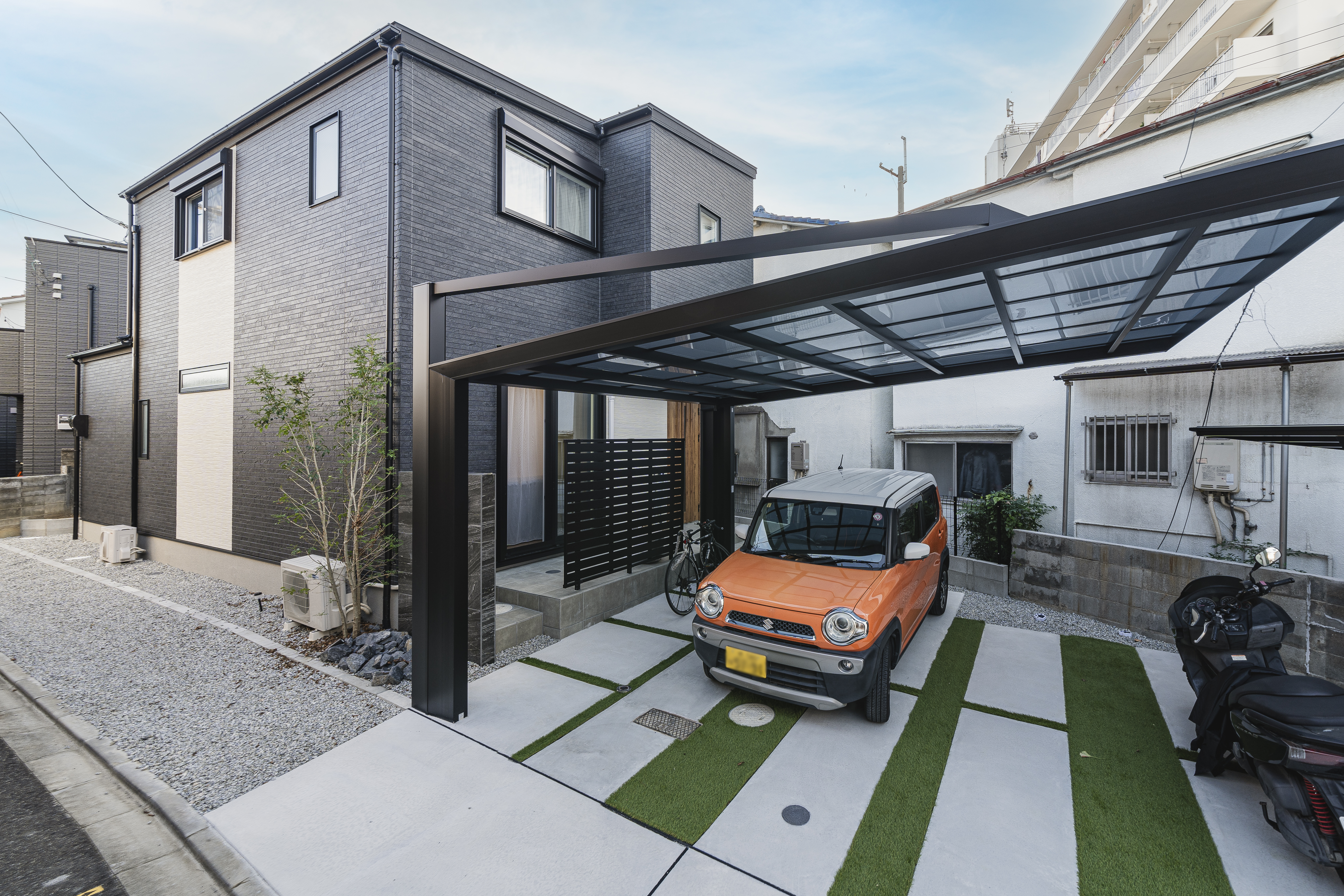 アーキホームライフ｜モダンなデザインの二階建て住宅の外観。ガレージには車が駐車されており、コンクリートとグリーンが調和した駐車スペースが印象的である。