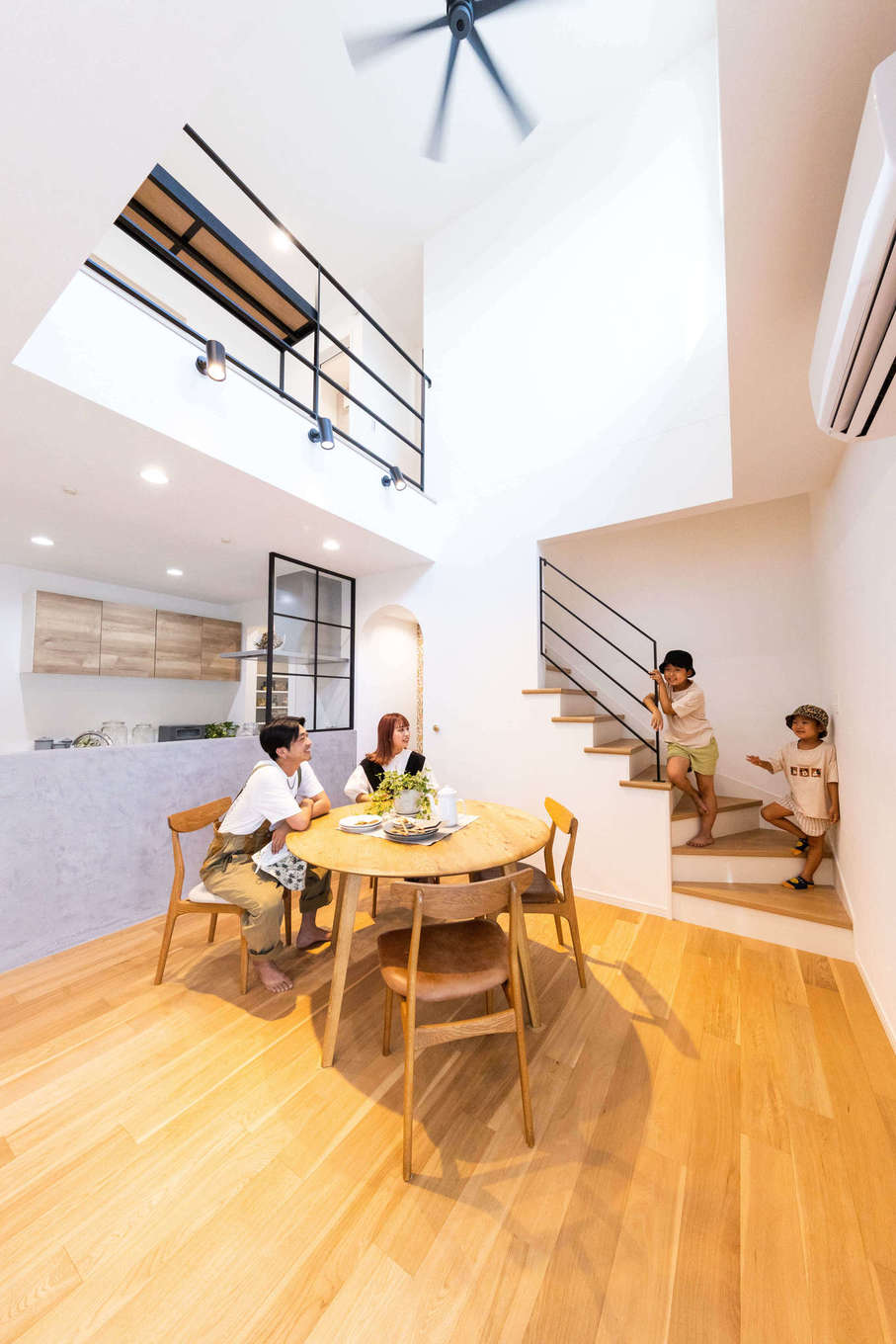 子育て安心住宅＆デザインラボ【デザイン住宅、趣味、インテリア】吹き抜けのダイニングスペース。モルタル仕上げのキッチン腰壁や、アイアンの階段の手すりなど、細部に夫妻のこだわりが反映されている。階段は玄関からLDKに入って一番奥に配置し、帰宅時にも外出時にも家族がいつでも自然と顔を合わせられるようにした