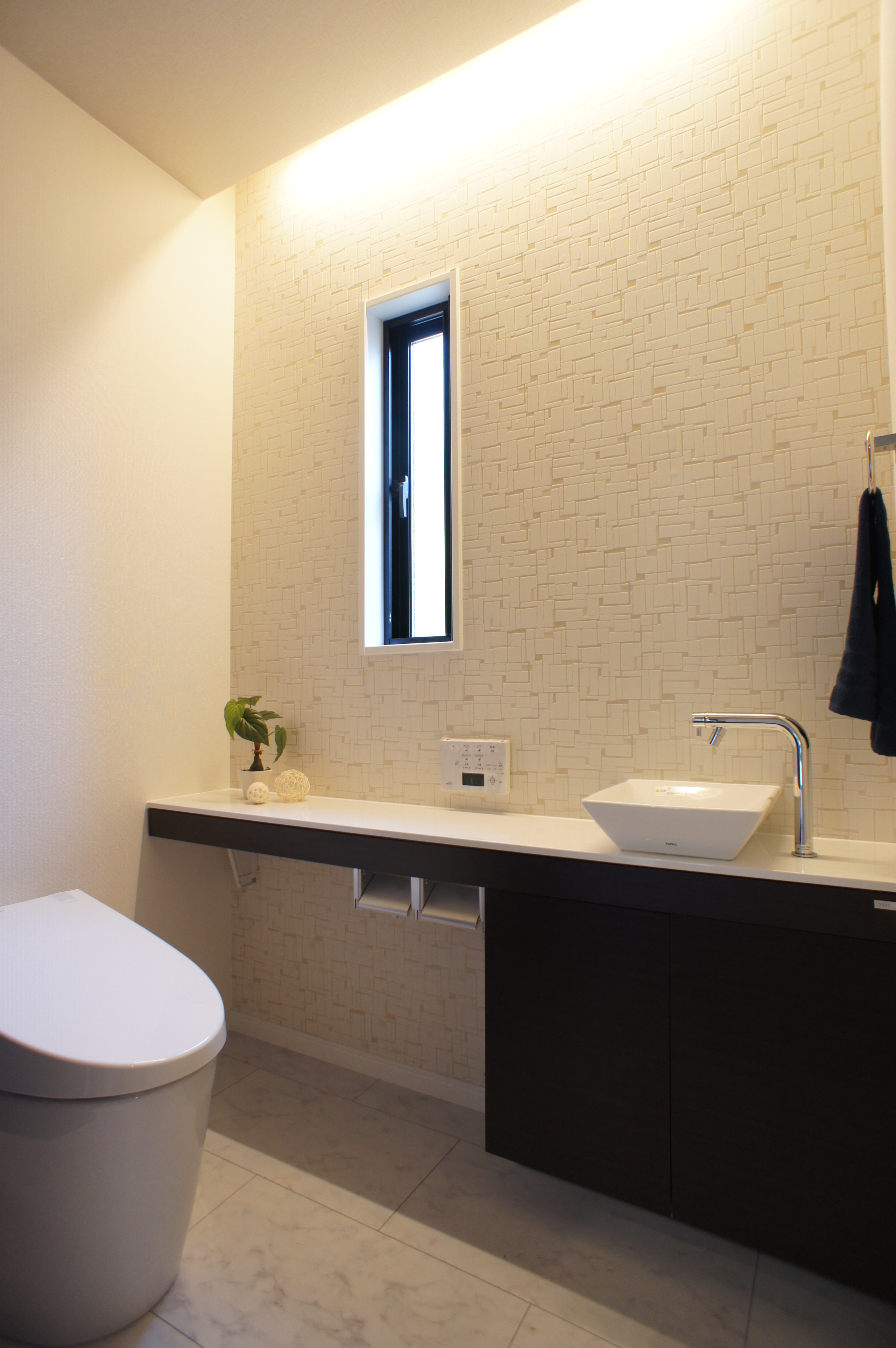 小野田産業～designers house project～｜落ち着いた色調のトイレ空間。洗練されたモダンなデザインで、壁の間接照明が柔らかな雰囲気を演出している。