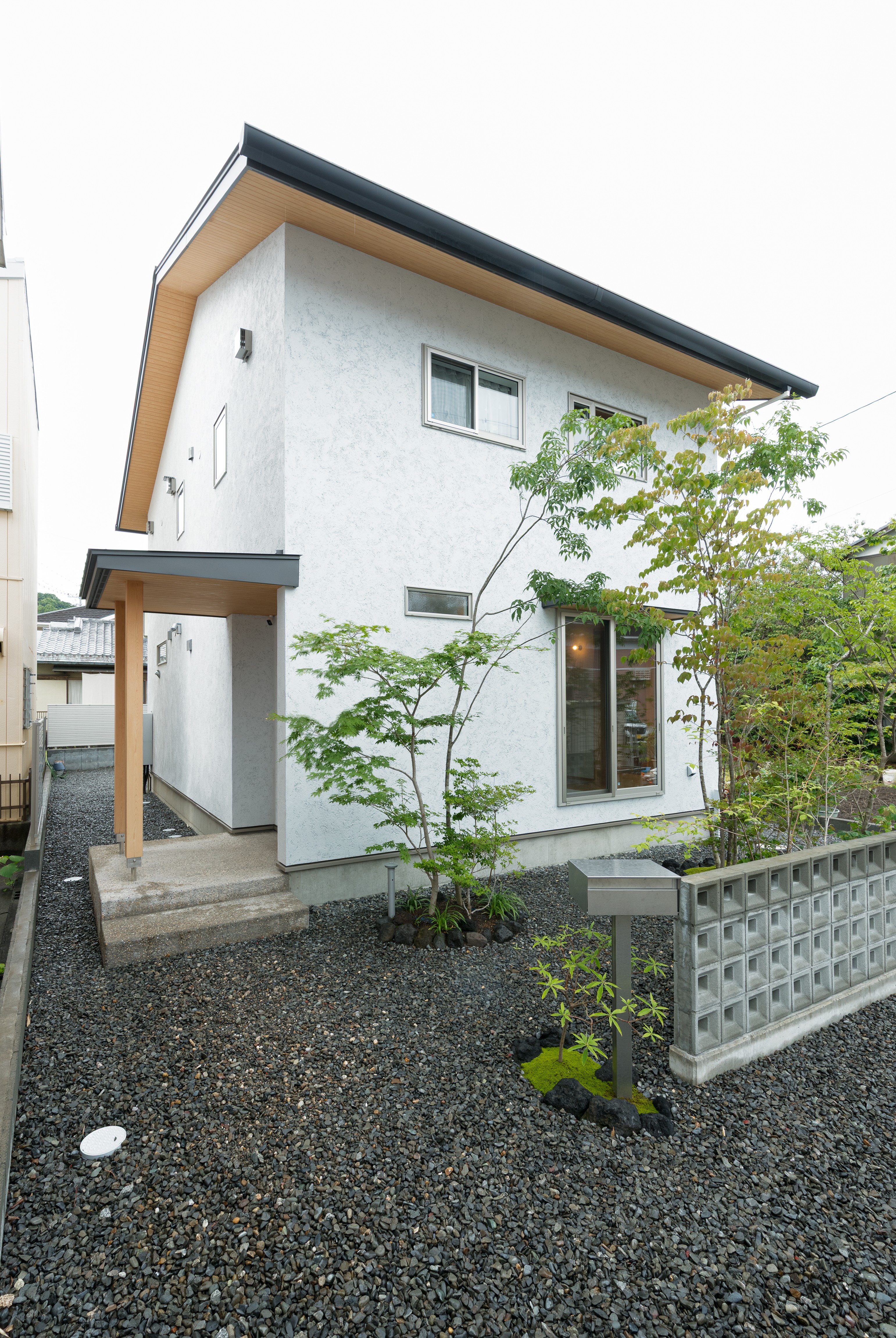 仲田工務店｜白いそとん壁の外観は、シンプルながらもどこか温かみを感じさせるデザイン。軒天の木目やアプローチの素材感が、自然と調和した上質な雰囲気を演出しています。玄関まわりや敷地内には落葉樹を中心に植栽を配置し、四季の変化を楽しめる庭に仕上げました。機能性と美しさを兼ね備えた、長く愛せる住まいです。