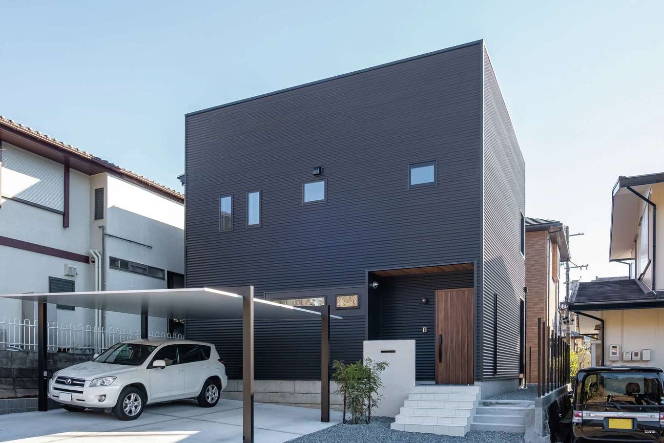 三和建設【デザイン住宅、子育て、間取り】黒ガルバリウムに玄関部分の軒天とドアの木目が映える。シンボルツリーは常緑樹のシマトネリコ
